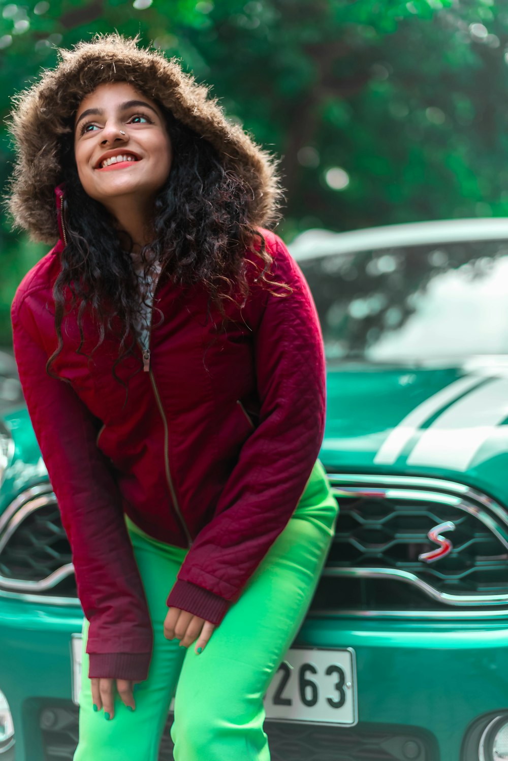 woman in red zip up jacket and green pants sitting on green car hood during daytime