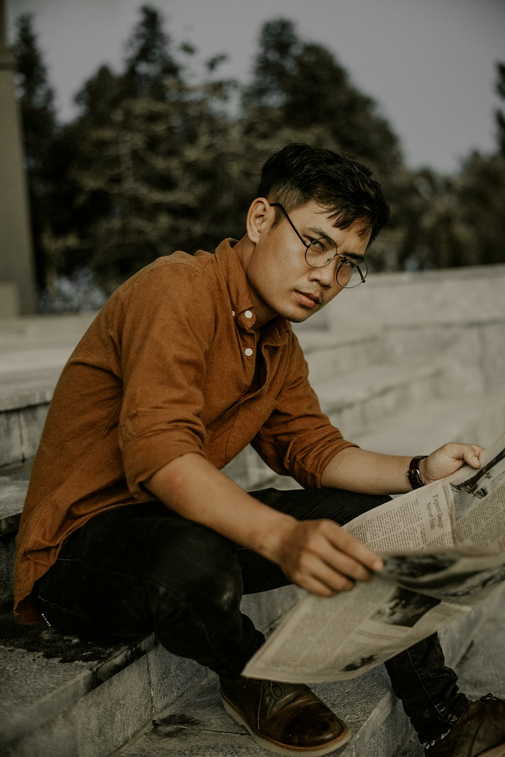 man in brown jacket and black pants sitting on concrete bench reading newspaper