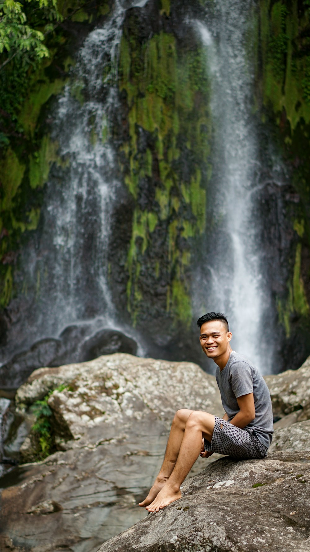 homme en t-shirt gris assis sur le rocher près des chutes d’eau pendant la journée