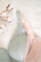 A close-up view of two delicate lace bras, one in soft green and the other in light pink, lying on a white surface. Beside them is a small branch with cotton flowers, adding a natural and cozy element to the composition.