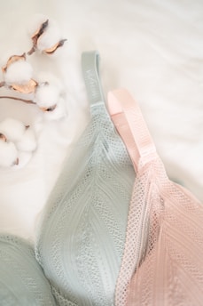 A close-up view of two delicate lace bras, one in soft green and the other in light pink, lying on a white surface. Beside them is a small branch with cotton flowers, adding a natural and cozy element to the composition.