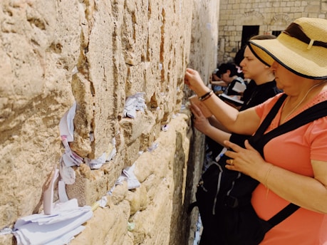People are praying at a stone wall, placing notes within the crevices. The wall is made of large, weathered stone blocks, and small pieces of paper are visible protruding from the gaps. The individuals appear focused and reverent.
