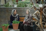 Two women are seated on a bench in a garden area, engaged in conversation. They are wearing headscarves and appear to be of different generations. In the foreground, there is a sculpture made from twisted tree branches with an attached bird figure. Two potted plants are placed near the bench, adding greenery to the scene. The background features a stone wall and some foliage.