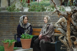 Two women are seated on a bench in a garden area, engaged in conversation. They are wearing headscarves and appear to be of different generations. In the foreground, there is a sculpture made from twisted tree branches with an attached bird figure. Two potted plants are placed near the bench, adding greenery to the scene. The background features a stone wall and some foliage.