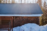 A wooden cabin with a metal roof surrounded by a snowy environment. A stack of chopped firewood is neatly arranged against the wall. Sunlight filters through the trees, casting a warm glow over the scene. A wooden swing is partially visible, suggesting a cozy spot for relaxation.