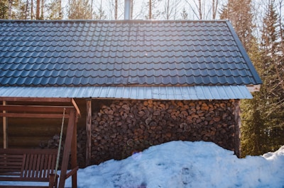 A wooden cabin with a metal roof surrounded by a snowy environment. A stack of chopped firewood is neatly arranged against the wall. Sunlight filters through the trees, casting a warm glow over the scene. A wooden swing is partially visible, suggesting a cozy spot for relaxation.
