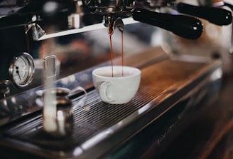 An espresso machine in use, with a cup placed beneath the spout collecting freshly brewed coffee. The metallic surface of the machine gleams with reflections. A small frothing pitcher is placed on the side, and there is a pressure gauge visible.