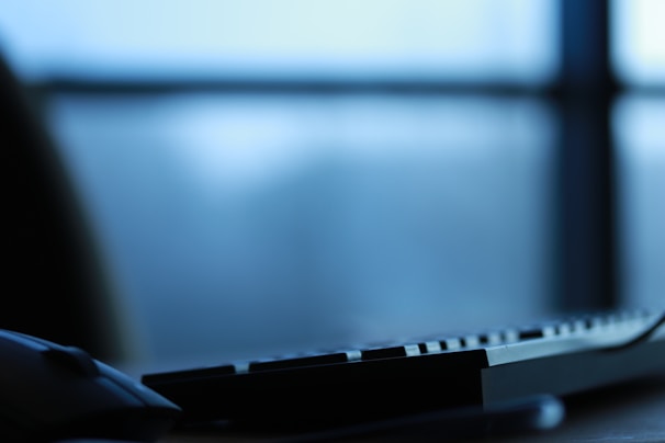 A close-up of a computer keyboard and mouse with a blurred window in the background. The lighting is soft and the image has a cool tone, suggesting early morning or late evening.