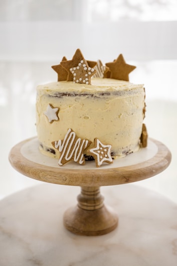 A decorated cake with light cream frosting sits atop a wooden cake stand. Adorned with star-shaped cookies and icing designs, the cake gives a whimsical and festive appearance. The background is soft and blurred, highlighting the cake's intricate details.