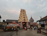A traditional Indian temple with an intricately carved gopuram stands in the center, surrounded by people walking on the paved pathway. The sky is overcast, adding a calming ambiance. Adjacent are structures housing a Tourist Information Center and a small shop. The scene includes a rath or chariot decorated with ornate details.