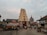 A traditional Indian temple with an intricately carved gopuram stands in the center, surrounded by people walking on the paved pathway. The sky is overcast, adding a calming ambiance. Adjacent are structures housing a Tourist Information Center and a small shop. The scene includes a rath or chariot decorated with ornate details.