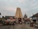 A traditional Indian temple with an intricately carved gopuram stands in the center, surrounded by people walking on the paved pathway. The sky is overcast, adding a calming ambiance. Adjacent are structures housing a Tourist Information Center and a small shop. The scene includes a rath or chariot decorated with ornate details.
