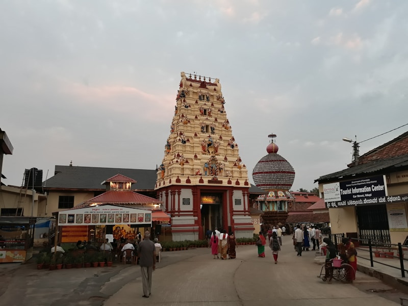 A traditional Indian temple with an intricately carved gopuram stands in the center, surrounded by people walking on the paved pathway. The sky is overcast, adding a calming ambiance. Adjacent are structures housing a Tourist Information Center and a small shop. The scene includes a rath or chariot decorated with ornate details.