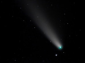 A bright comet with a glowing nucleus and a long tail streaks across a star-filled night sky. The tail is illuminated and tapers off into the darkness, while the stars remain as small, scattered points of light.