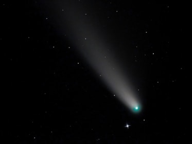 A bright comet with a glowing nucleus and a long tail streaks across a star-filled night sky. The tail is illuminated and tapers off into the darkness, while the stars remain as small, scattered points of light.