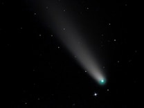 A bright comet with a glowing nucleus and a long tail streaks across a star-filled night sky. The tail is illuminated and tapers off into the darkness, while the stars remain as small, scattered points of light.