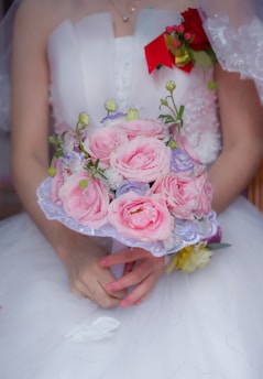 A bride holds a delicate bouquet of pink roses accented with small white and purple flowers. The bride's elegant white dress features detailed lace patterns, and there is a vibrant red and yellow corsage pinned to the front. Her hands, with neatly manicured nails, gently cradle the bouquet.