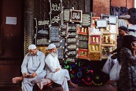 Two men dressed in traditional attire are sitting near a wall adorned with framed Islamic calligraphy art. One man is looking at a phone, while the other is looking ahead. Behind them, various ornate frames display religious inscriptions. Other people are seen walking by, each carrying plastic bags.