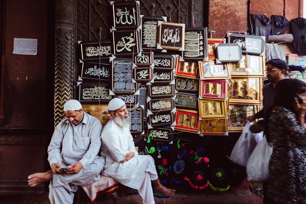 Two men dressed in traditional attire are sitting near a wall adorned with framed Islamic calligraphy art. One man is looking at a phone, while the other is looking ahead. Behind them, various ornate frames display religious inscriptions. Other people are seen walking by, each carrying plastic bags.