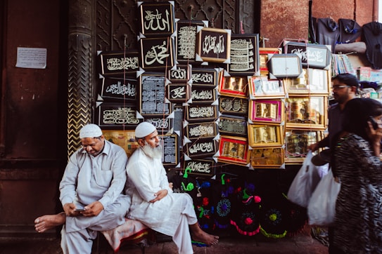 Two men dressed in traditional attire are sitting near a wall adorned with framed Islamic calligraphy art. One man is looking at a phone, while the other is looking ahead. Behind them, various ornate frames display religious inscriptions. Other people are seen walking by, each carrying plastic bags.