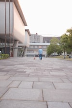 A person in casual attire is walking a dog on a wide paved walkway, lined with modern architectural buildings. The setting appears to be an urban or campus area with large buildings and greenery in the background.