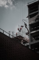 Construction workers wearing safety gear are working at a high altitude on a building structure. A crane is lifting materials, and the building frame is partially complete with visible steel beams and netting for safety.