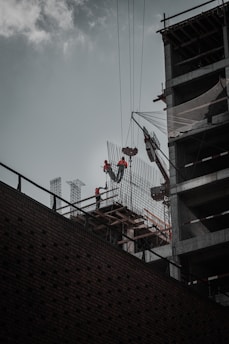 Construction workers wearing safety gear are working at a high altitude on a building structure. A crane is lifting materials, and the building frame is partially complete with visible steel beams and netting for safety.