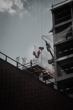 Construction workers wearing safety gear are working at a high altitude on a building structure. A crane is lifting materials, and the building frame is partially complete with visible steel beams and netting for safety.