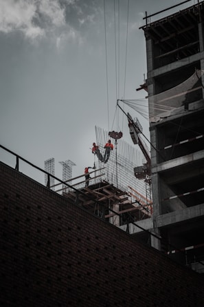 Construction workers wearing safety gear are working at a high altitude on a building structure. A crane is lifting materials, and the building frame is partially complete with visible steel beams and netting for safety.