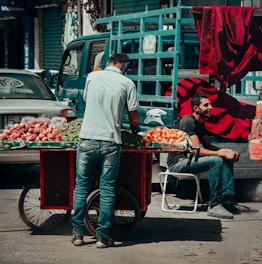 A street vendor with a cart full of various fruits such as tomatoes and possibly apples. One man is standing and arranging the produce while another man is sitting nearby on a chair, resting. The background features a parked blue truck and a car with a license plate written in Arabic. Bright red fabric is draped over the truck.