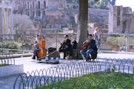 A group of street musicians is performing outdoors near ancient ruins. The band includes a double bassist and a cellist, with a music stand and some equipment at their feet. A few people are watching the performance, while the musicians are seated near a tree, and the background is filled with historic stone structures and lush greenery.