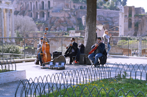 A group of street musicians is performing outdoors near ancient ruins. The band includes a double bassist and a cellist, with a music stand and some equipment at their feet. A few people are watching the performance, while the musicians are seated near a tree, and the background is filled with historic stone structures and lush greenery.