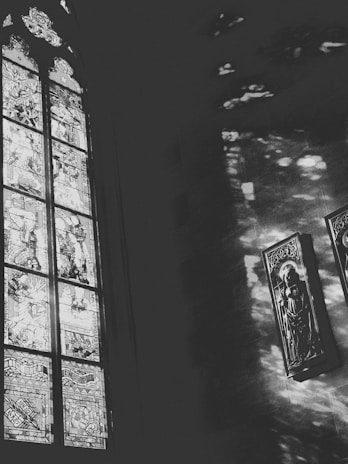 A black and white image featuring a large stained glass window on the left, depicting intricate scenes with human figures. Light filters through the glass, casting patterns on the adjacent wall. Two framed religious artworks hang on the right, likely depicting saints.