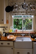A cozy kitchen scene with a window overlooking a lush green garden, adorned with floral-patterned curtains. Above the window, an array of pots and pans hang neatly. The sink area is tidy with washing liquid, a couple of mugs, and a bowl containing kitchen utensils. The countertop has a warm, wooden finish, complemented by decorative tiles.