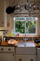 A cozy kitchen scene with a window overlooking a lush green garden, adorned with floral-patterned curtains. Above the window, an array of pots and pans hang neatly. The sink area is tidy with washing liquid, a couple of mugs, and a bowl containing kitchen utensils. The countertop has a warm, wooden finish, complemented by decorative tiles.