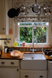 A cozy kitchen scene with a window overlooking a lush green garden, adorned with floral-patterned curtains. Above the window, an array of pots and pans hang neatly. The sink area is tidy with washing liquid, a couple of mugs, and a bowl containing kitchen utensils. The countertop has a warm, wooden finish, complemented by decorative tiles.