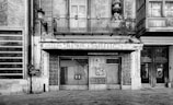 A black and white photograph of an old storefront with ornate architectural details. The facade features a balcony with iron railings, tiled designs, and a weathered sign that reads 'Azulejos Ramon Castello Pavimentos.' The entrance includes a decorative wall with a mural and a closed gate.