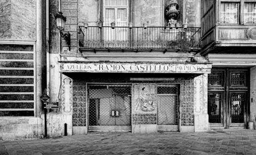 A black and white photograph of an old storefront with ornate architectural details. The facade features a balcony with iron railings, tiled designs, and a weathered sign that reads 'Azulejos Ramon Castello Pavimentos.' The entrance includes a decorative wall with a mural and a closed gate.