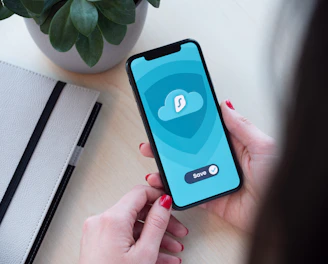 A person holding a smartphone displaying an app with a cloud icon and a save button. The background includes a potted plant and a closed notebook on a light-colored desk.