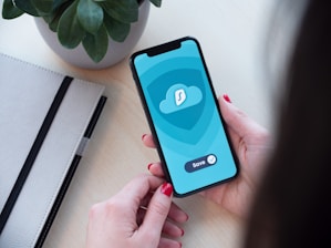 A person holding a smartphone displaying an app with a cloud icon and a save button. The background includes a potted plant and a closed notebook on a light-colored desk.