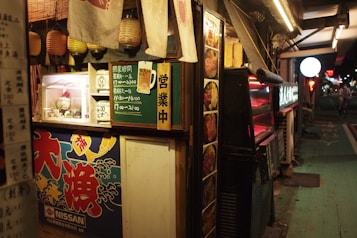 A small, street-side food stall featuring Japanese signage and decorations. Hanging lanterns and cloth banners are visible above the counter area, which has images of food displayed. A chalkboard lists operating hours, and there are bright, colorful illustrations and signs in Japanese characters around the stall. A bicycle and streetlights can be seen in the background, suggesting an urban nighttime setting.
