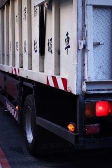A segment of a large vehicle with a metal structure, featuring black painted characters on the side, likely indicating a function or purpose. The rear section includes red and amber lights, with diagonal red and white striped patterns adding visibility. Part of a tire and metal framing are visible, suggesting a truck or similar heavy-duty vehicle.