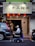 A person in a mobility scooter is moving past a storefront with a sign featuring Chinese characters. Several red lanterns are hanging under the sign, illuminating the entrance. Parked vehicles, including a car and a motorcycle, are present on the street beside the storefront.