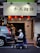 A person in a mobility scooter is moving past a storefront with a sign featuring Chinese characters. Several red lanterns are hanging under the sign, illuminating the entrance. Parked vehicles, including a car and a motorcycle, are present on the street beside the storefront.