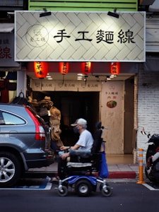 A person in a mobility scooter is moving past a storefront with a sign featuring Chinese characters. Several red lanterns are hanging under the sign, illuminating the entrance. Parked vehicles, including a car and a motorcycle, are present on the street beside the storefront.