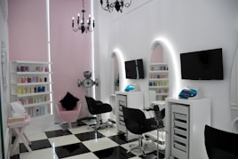 A modern salon with sleek black and white checkered flooring and a minimalist aesthetic. The room features two black salon chairs facing stylish white stations with illuminated mirrors and mounted televisions. A variety of hair products are neatly arranged on shelves, and a chandelier adds an elegant touch. The walls are a soft pink, complementing the contemporary decor.