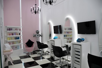A modern salon with sleek black and white checkered flooring and a minimalist aesthetic. The room features two black salon chairs facing stylish white stations with illuminated mirrors and mounted televisions. A variety of hair products are neatly arranged on shelves, and a chandelier adds an elegant touch. The walls are a soft pink, complementing the contemporary decor.