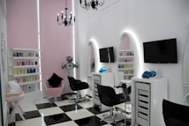A modern salon with sleek black and white checkered flooring and a minimalist aesthetic. The room features two black salon chairs facing stylish white stations with illuminated mirrors and mounted televisions. A variety of hair products are neatly arranged on shelves, and a chandelier adds an elegant touch. The walls are a soft pink, complementing the contemporary decor.