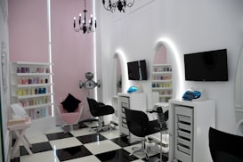 A modern salon with sleek black and white checkered flooring and a minimalist aesthetic. The room features two black salon chairs facing stylish white stations with illuminated mirrors and mounted televisions. A variety of hair products are neatly arranged on shelves, and a chandelier adds an elegant touch. The walls are a soft pink, complementing the contemporary decor.