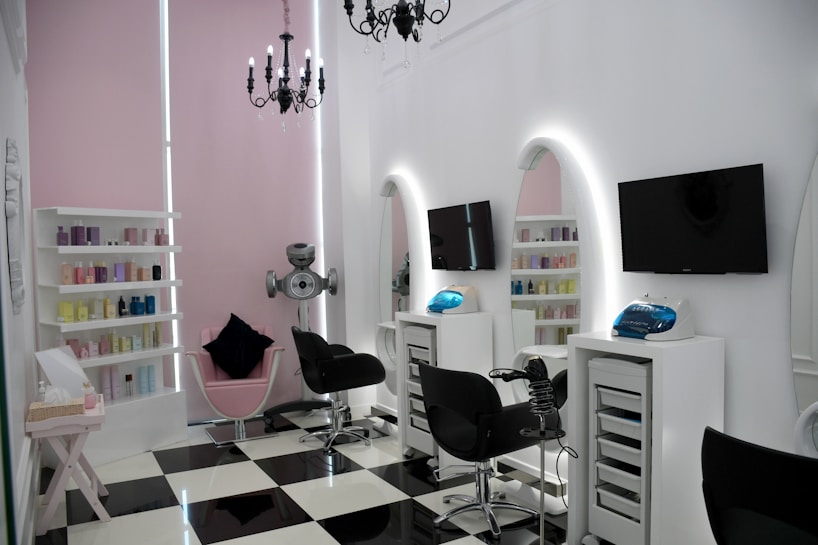 A modern salon with sleek black and white checkered flooring and a minimalist aesthetic. The room features two black salon chairs facing stylish white stations with illuminated mirrors and mounted televisions. A variety of hair products are neatly arranged on shelves, and a chandelier adds an elegant touch. The walls are a soft pink, complementing the contemporary decor.